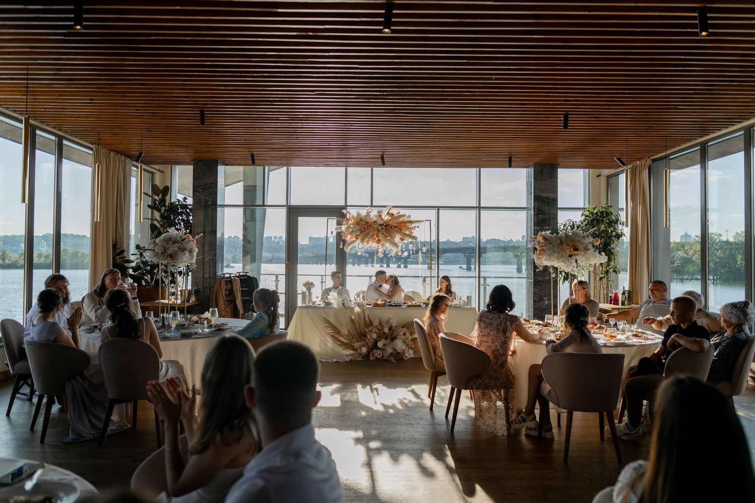 Elegant waterfront wedding reception with guests dining at decorated tables, large windows overlooking a lake, wood ceiling.