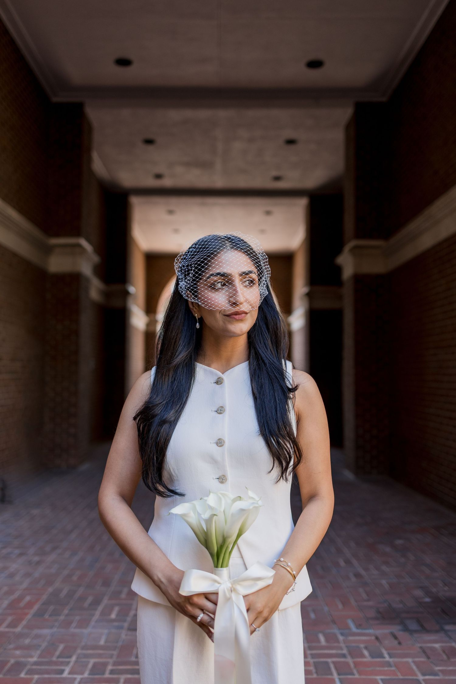 Bride in white sleeveless gown and birdcage veil holds calla lily bouquet with ribbon in brick hallway.