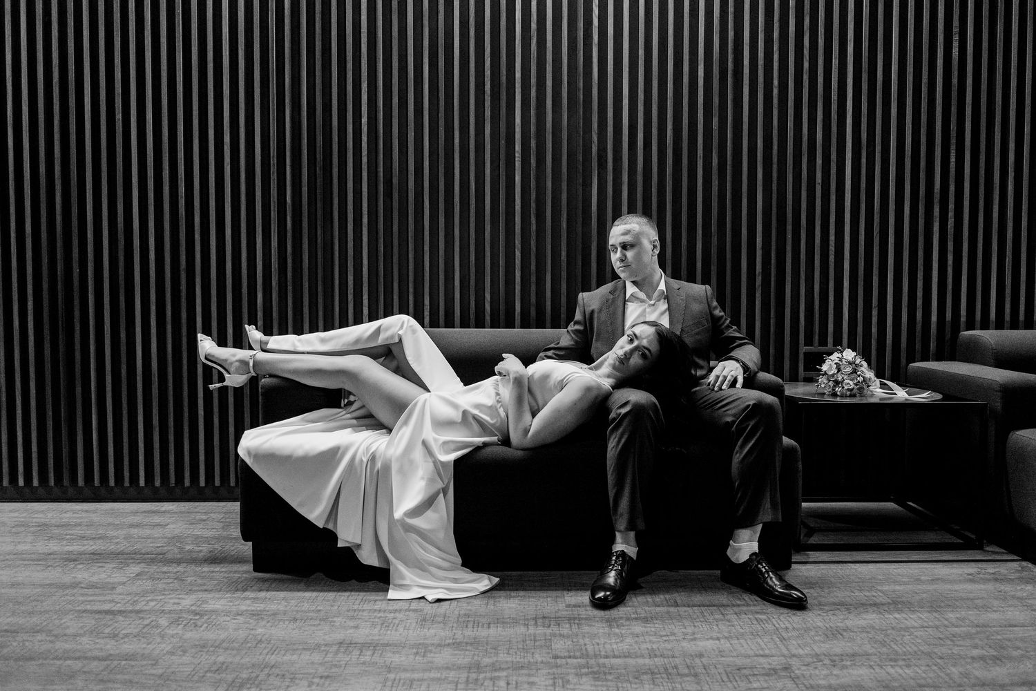 Black and white photo of bride reclining across groom's lap on a sofa, with a bouquet nearby.