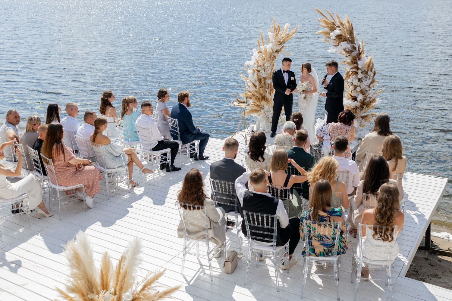 Outdoor waterfront wedding ceremony on white dock with floral arch, guests seated in white chairs, bride and groom at altar.