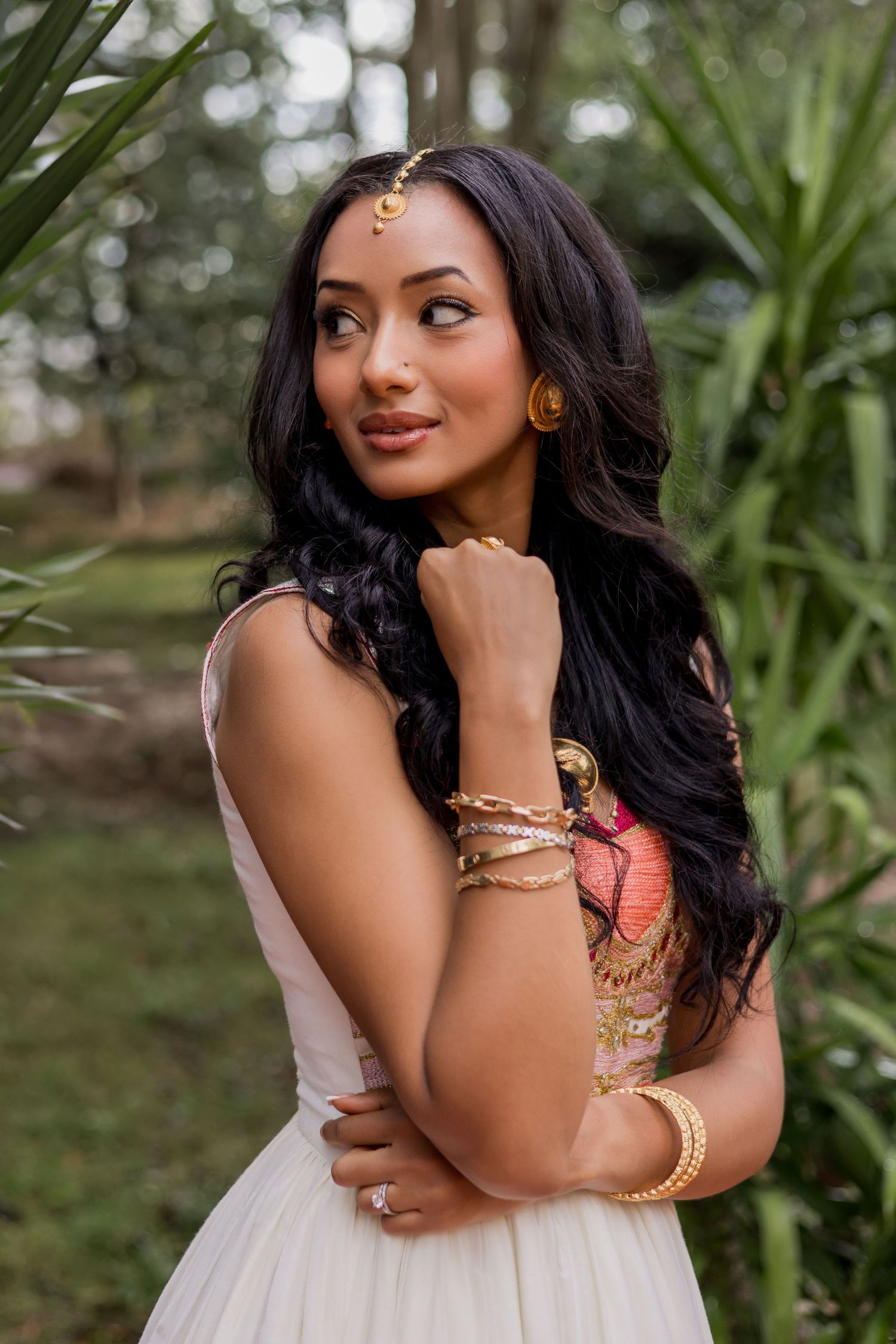 Young woman in white dress with gold jewelry and maang tikka poses gracefully outdoors among lush green foliage.