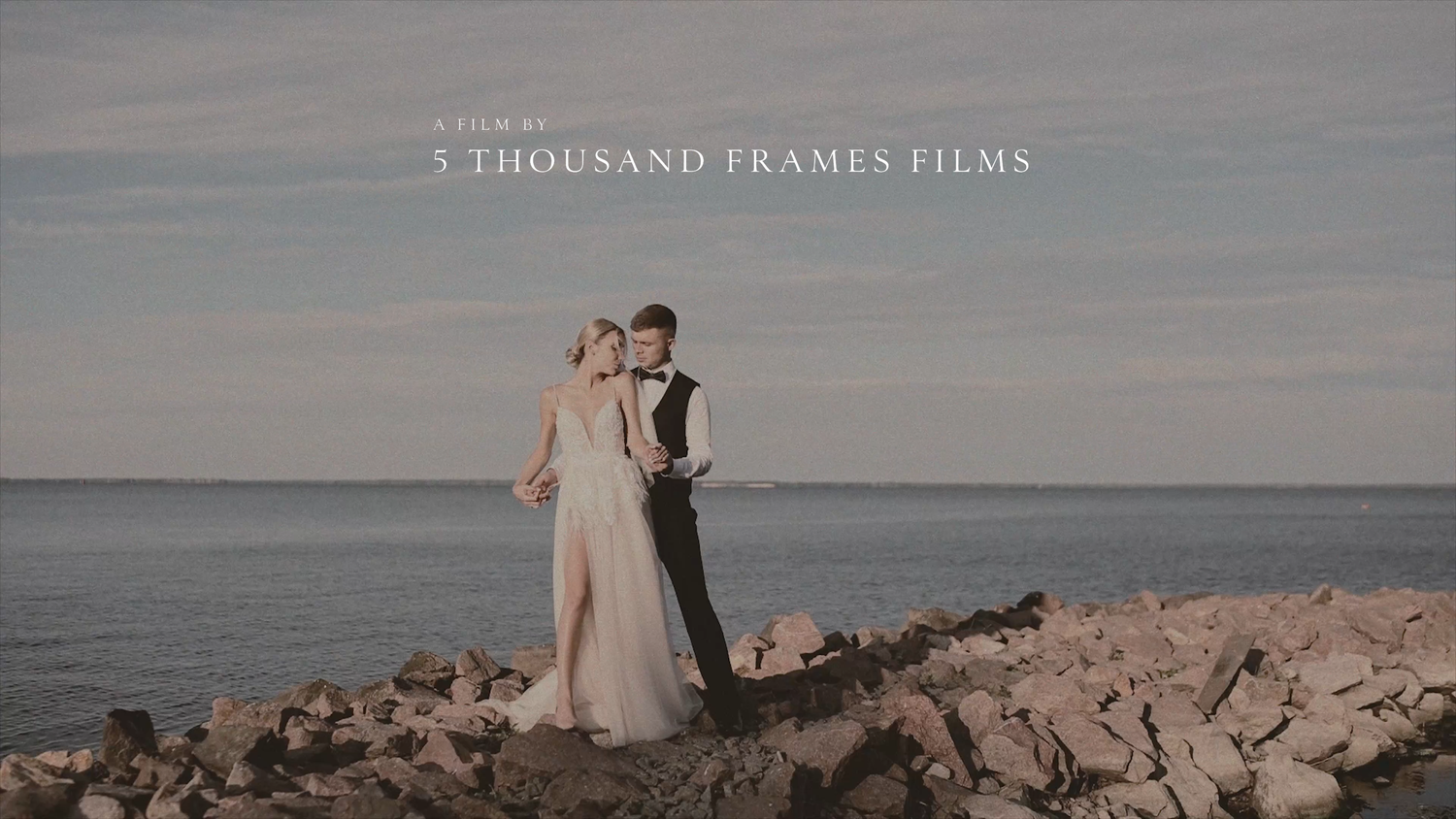 A bride and groom stand romantically on rocky coastal cliffs by the sea, filmed by 5 Thousand Frames Films.