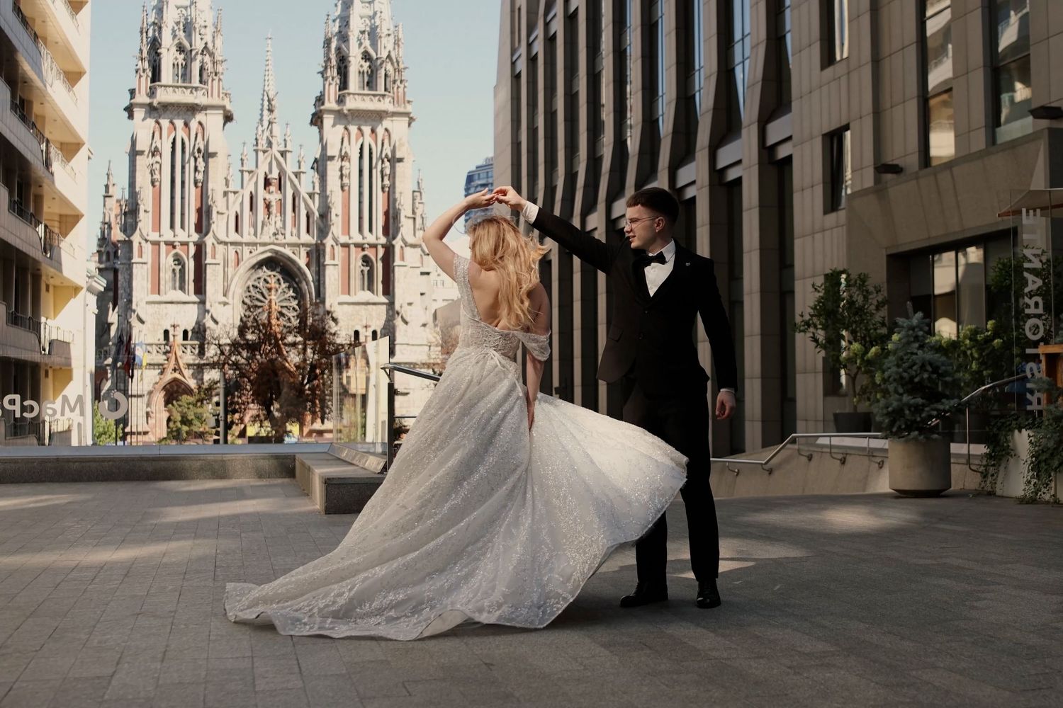 Bride in white gown and groom in tuxedo dancing outdoors near a Gothic cathedral backdrop.