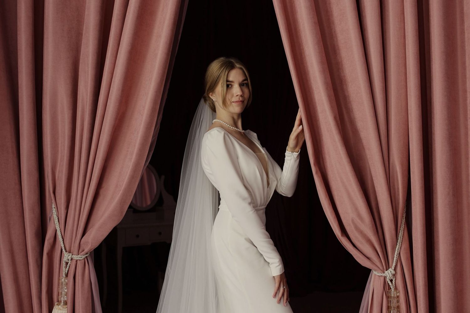 Elegant bride in white gown and veil standing between dusty rose velvet curtains, smiling gracefully.