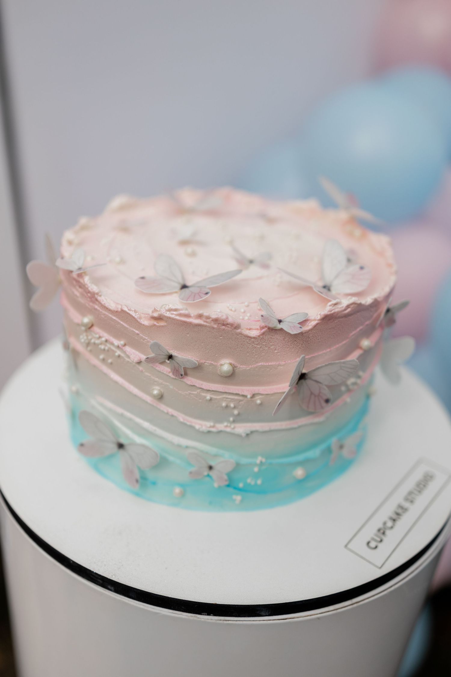 A pink and blue ombre cake decorated with white butterflies on a white pedestal.