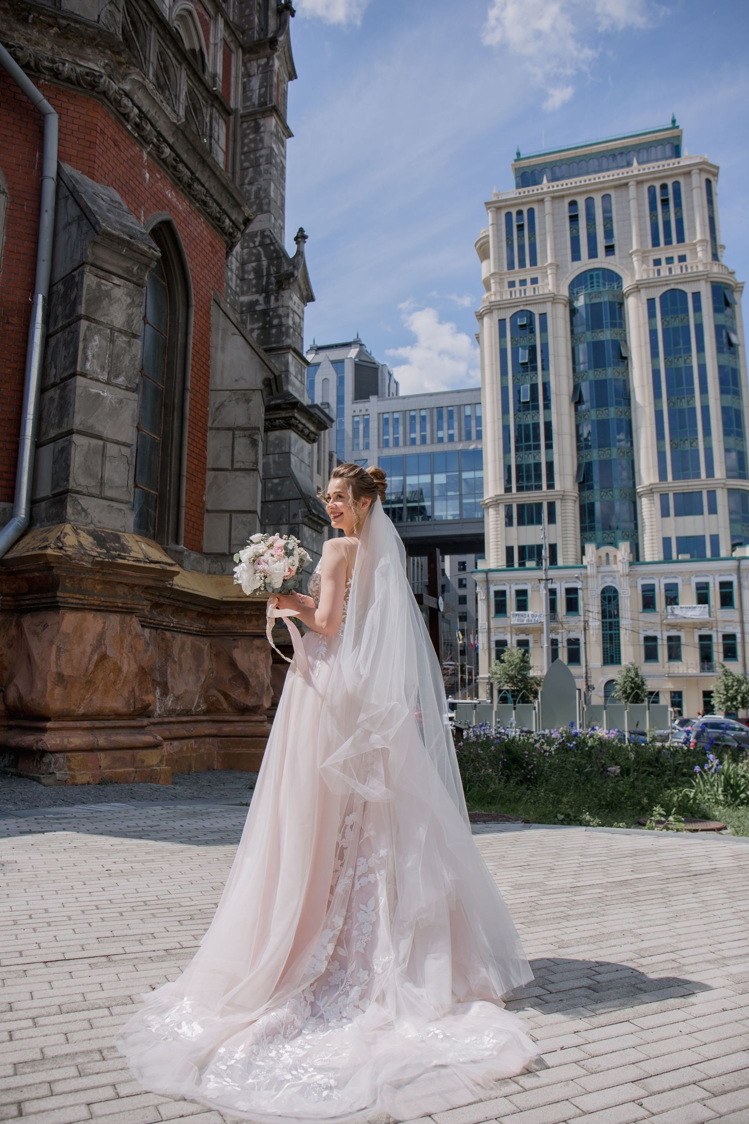 Elegant bride in lace gown with veil holds bouquet outside historic church with modern skyscraper backdrop.