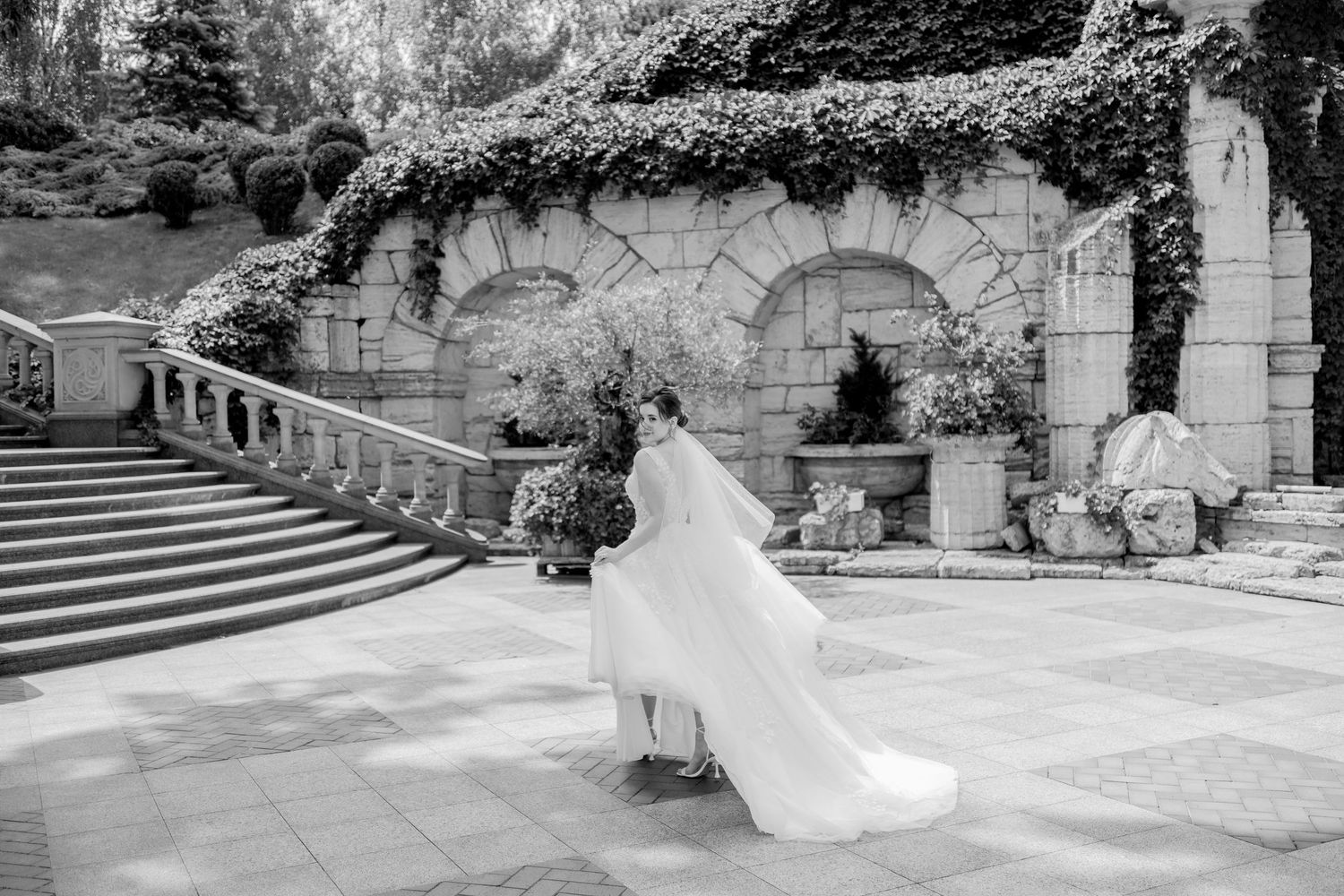Black and white photo of a bride in a flowing gown and veil walking through an elegant stone garden venue.
