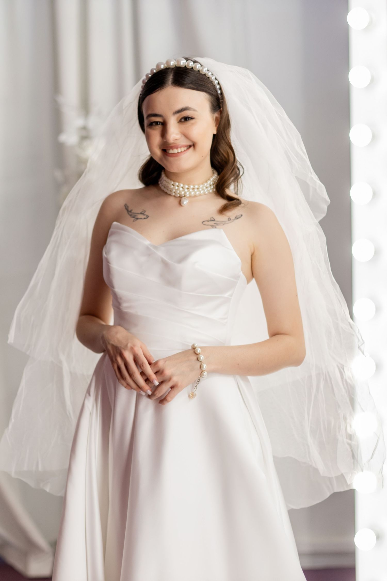 Smiling bride in strapless white gown, pearl jewelry, and veil poses elegantly in a bright bridal studio.