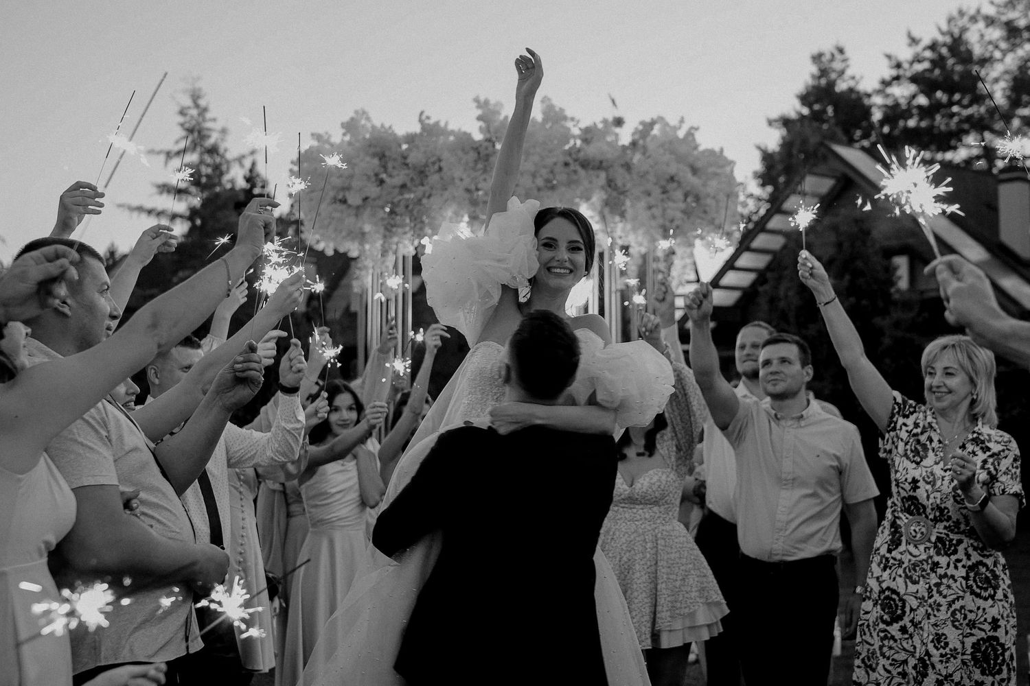 Black and white wedding photo of bride being lifted by groom surrounded by guests holding sparklers outdoors.