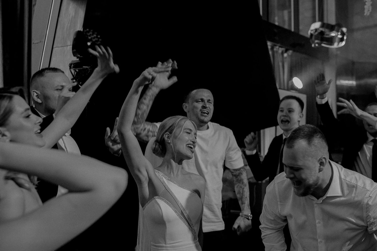 Black and white wedding photo of bride and guests celebrating joyfully, raising hands and dancing at reception.