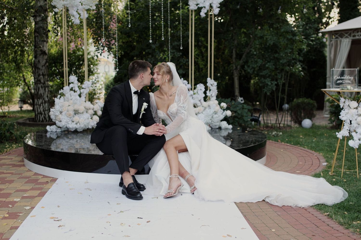 A bride and groom share a romantic moment on a white aisle at an elegant outdoor wedding ceremony with floral decor.