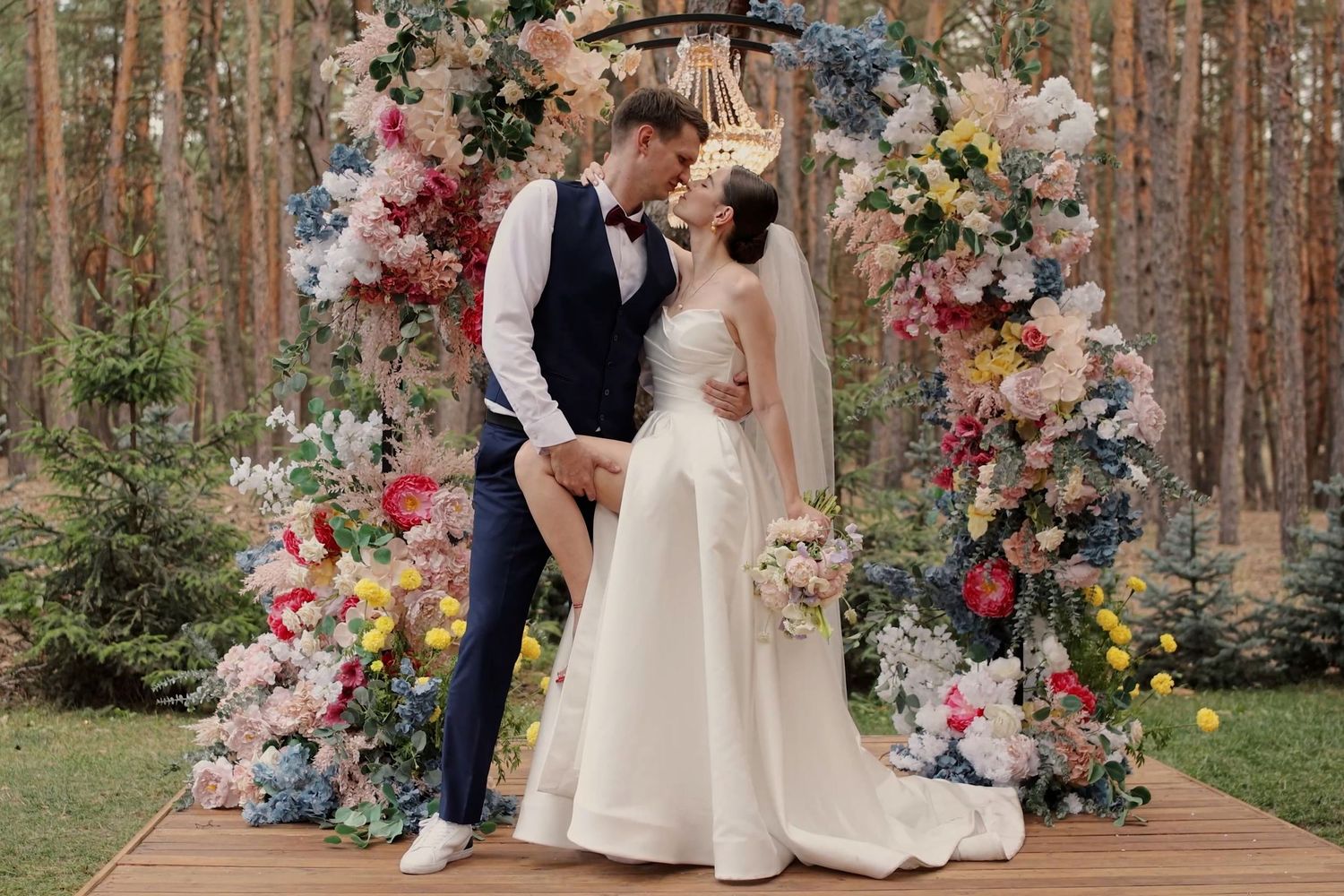 Bride and groom kissing at outdoor wedding altar adorned with colorful floral arch in a forest setting.