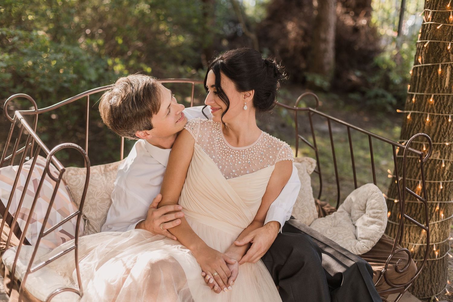 Bride in lace wedding gown sitting on ornate bench with young boy, sharing a joyful moment outdoors.