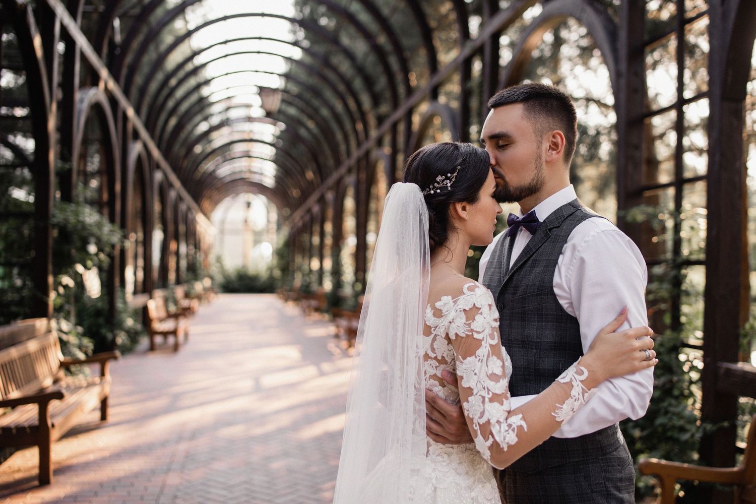 A bride and groom share a romantic kiss in an elegant arched greenhouse corridor on their wedding day.