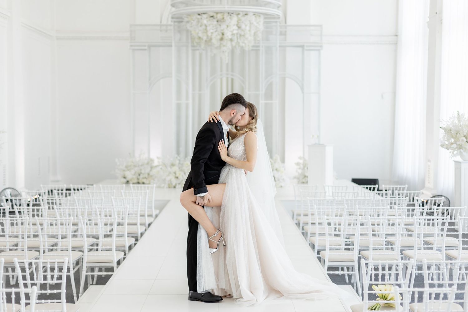 Bride and groom share romantic embrace at elegant all-white wedding ceremony venue with floral decor and ghost chairs.