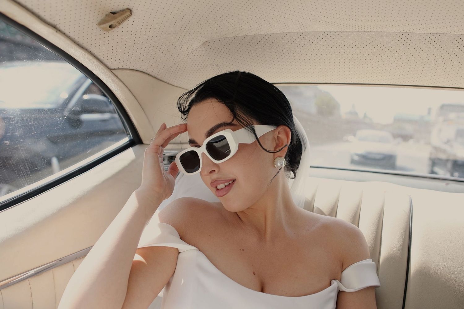 A stylish woman in a white off-shoulder top wearing white sunglasses poses in a vintage car interior.
