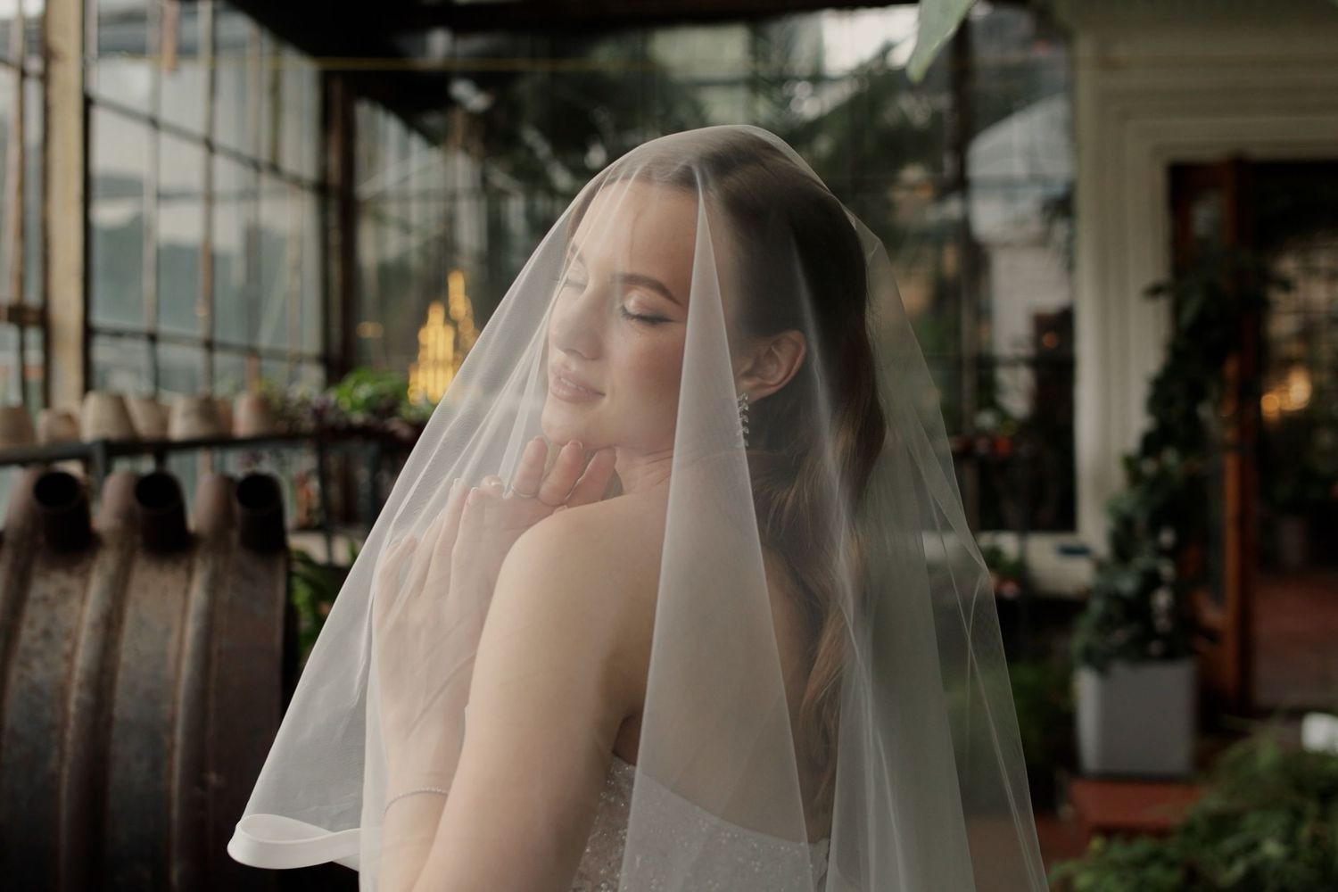 Bride in white gown with sheer veil draped over head, posing elegantly in a lush greenhouse setting.