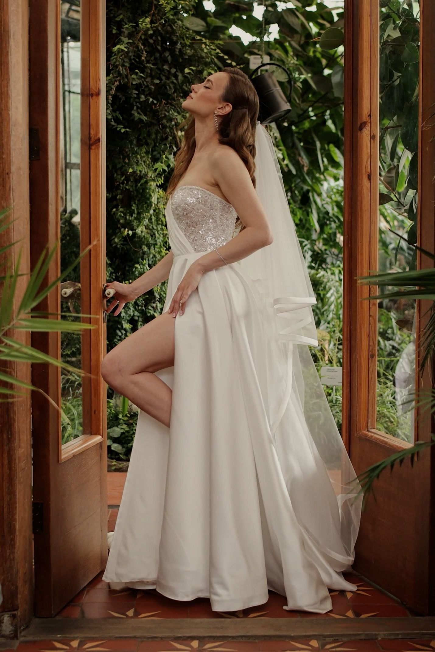 Bride in strapless sequin bodice wedding gown with high slit and veil poses in greenhouse doorway.