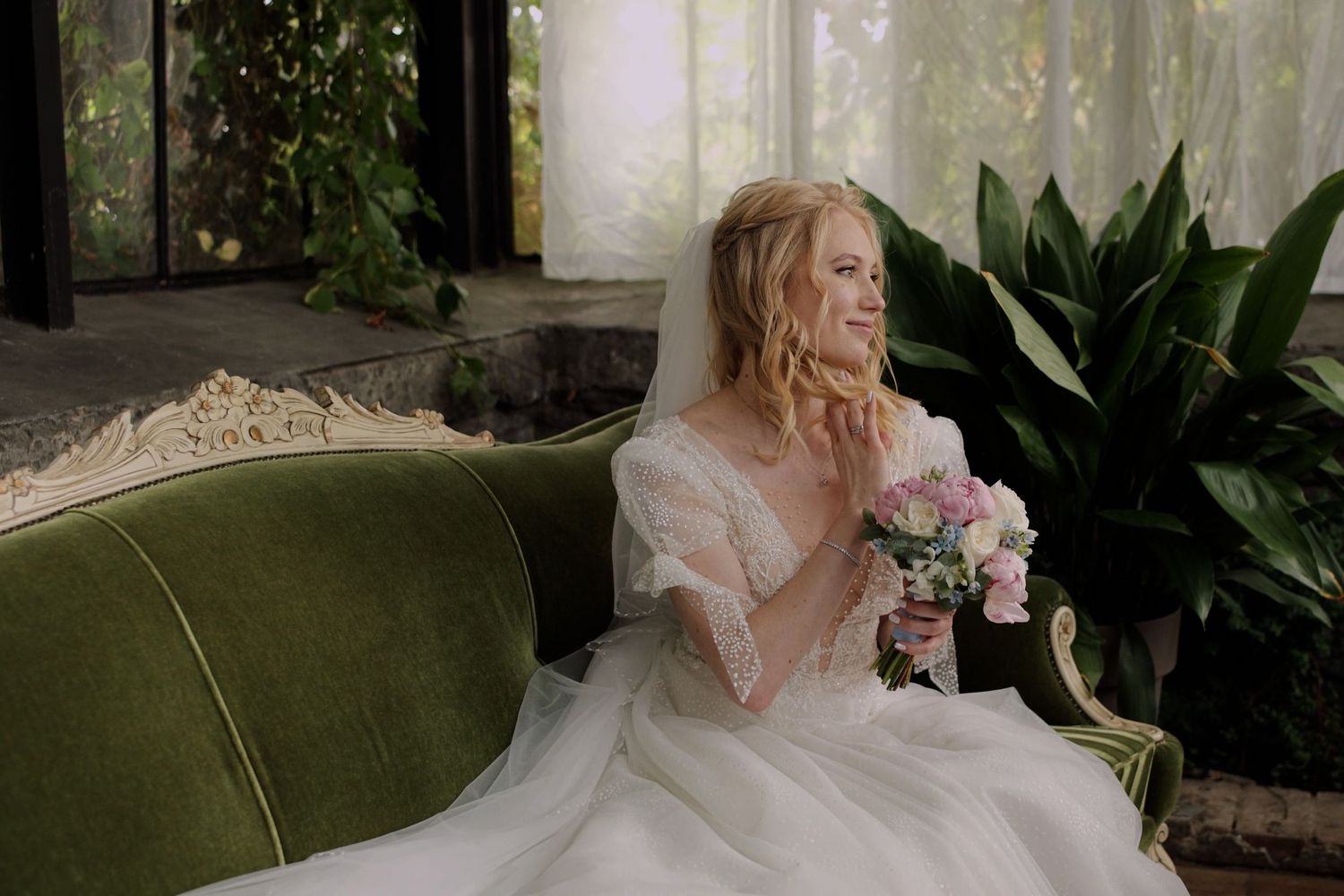 Bride in white gown holding pink bouquet, seated on green velvet sofa, smiling in elegant garden setting.