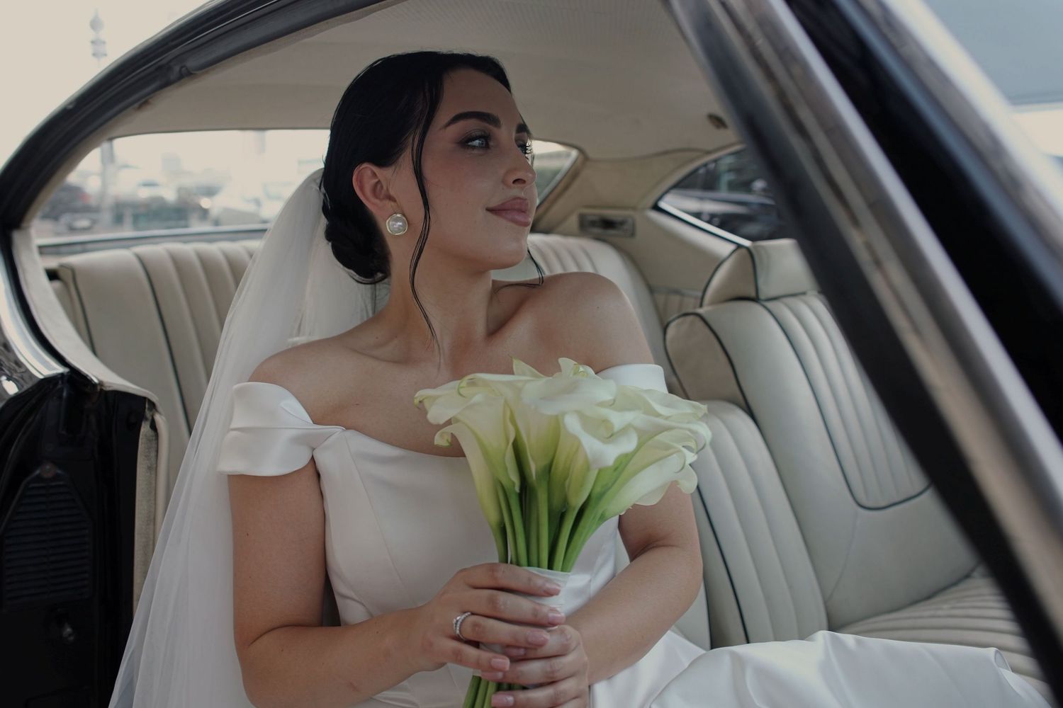 Bride in white off-shoulder gown holding calla lily bouquet sitting in luxury car interior on wedding day.