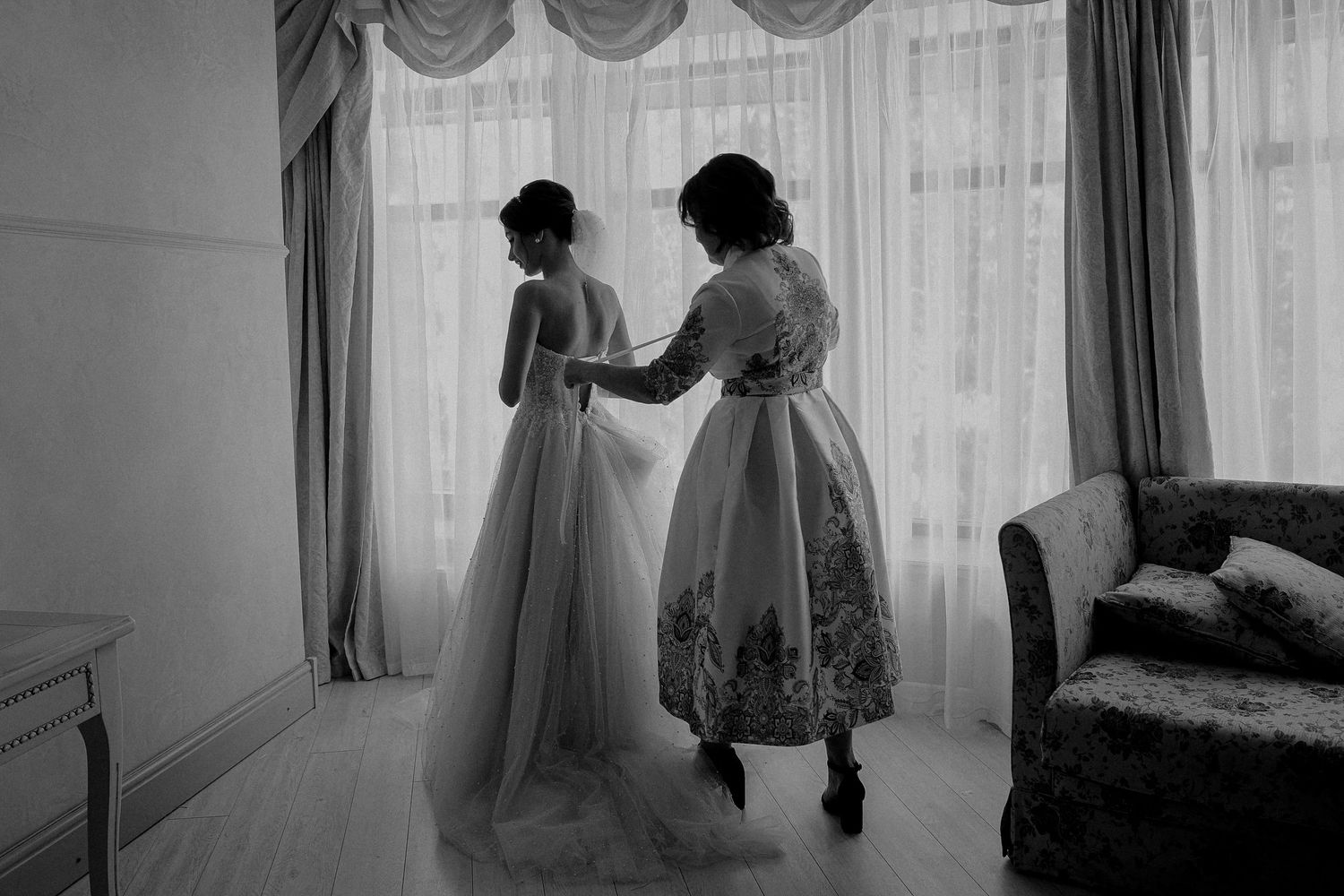 Black and white photo of bride getting dress fastened by helper near bright window in elegant room.