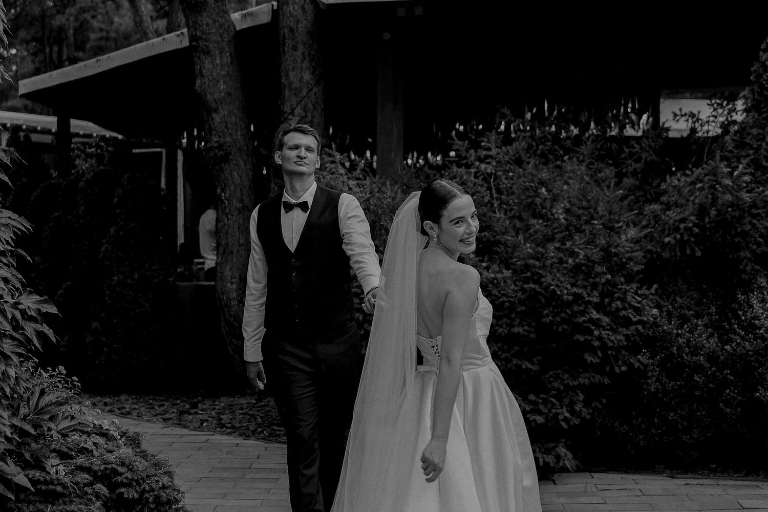 Black and white photo of a bride and groom walking hand in hand through a lush garden on their wedding day.