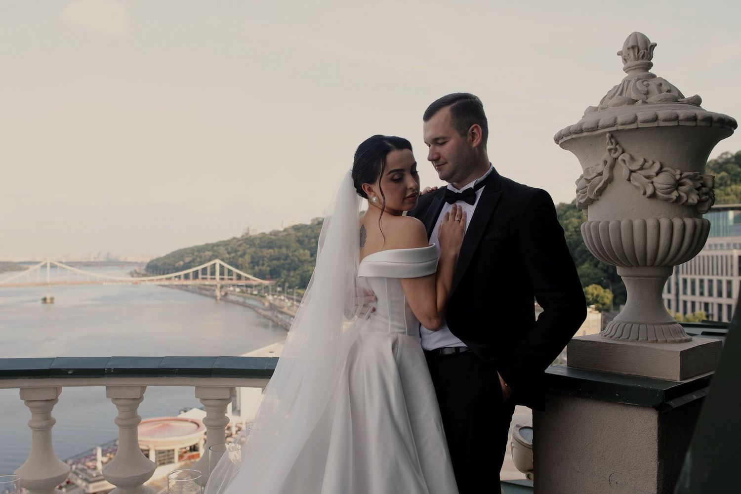A bride and groom share a romantic moment on a balcony overlooking a river and bridge on their wedding day.