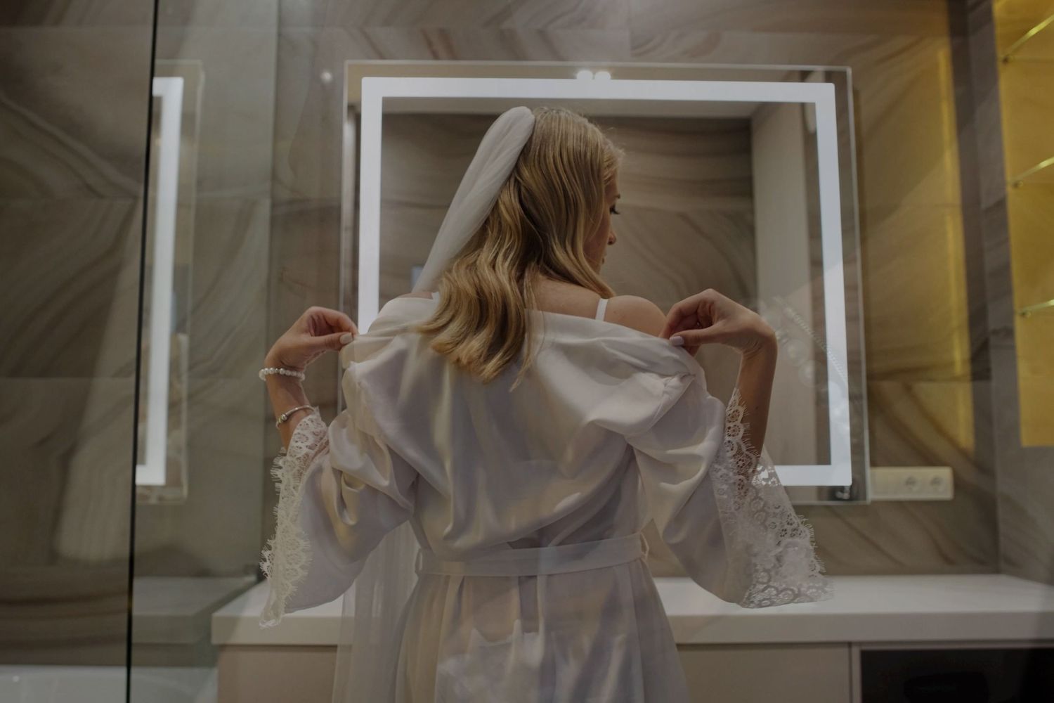 Bride in white robe adjusting veil in elegant bathroom mirror, wedding morning preparation.
