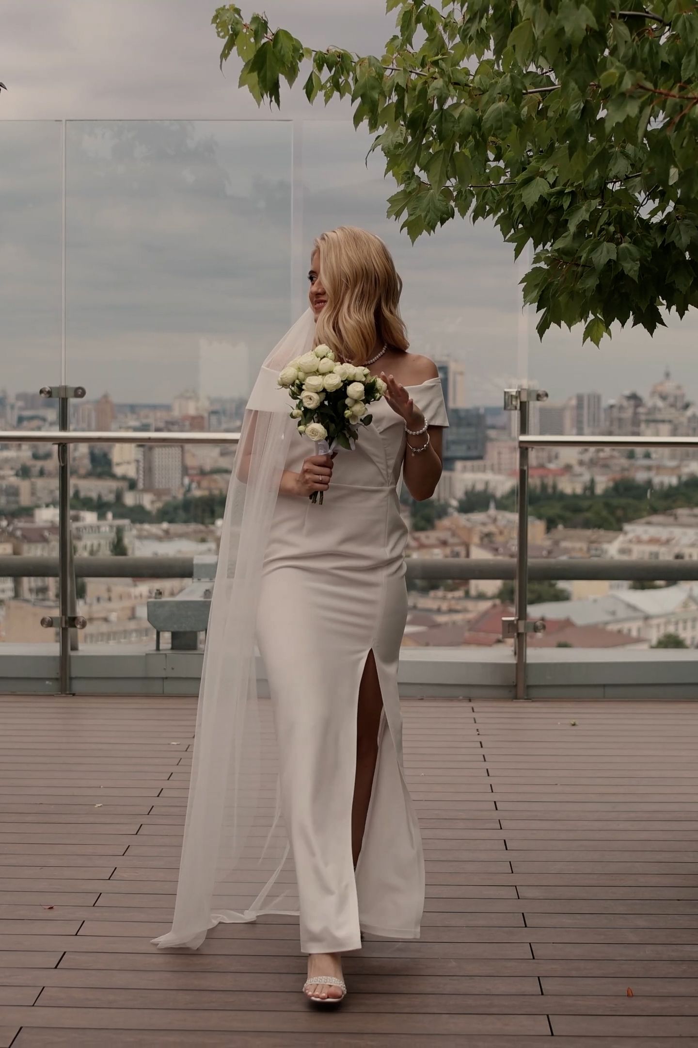 Elegant bride in white gown and veil holds bouquet on rooftop terrace with city skyline backdrop.