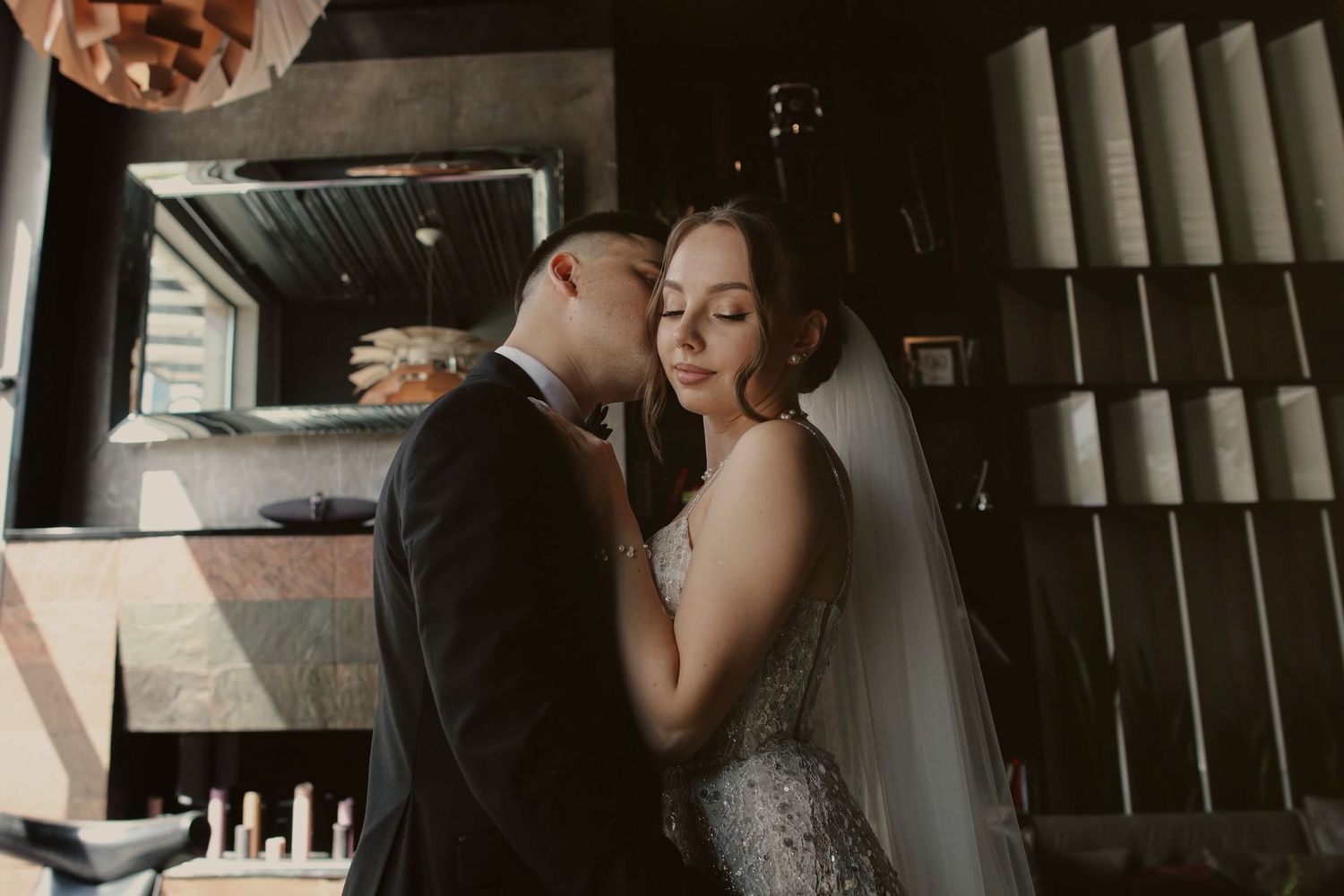 A bride and groom share an intimate moment together indoors, with the bride wearing a veil and elegant gown.