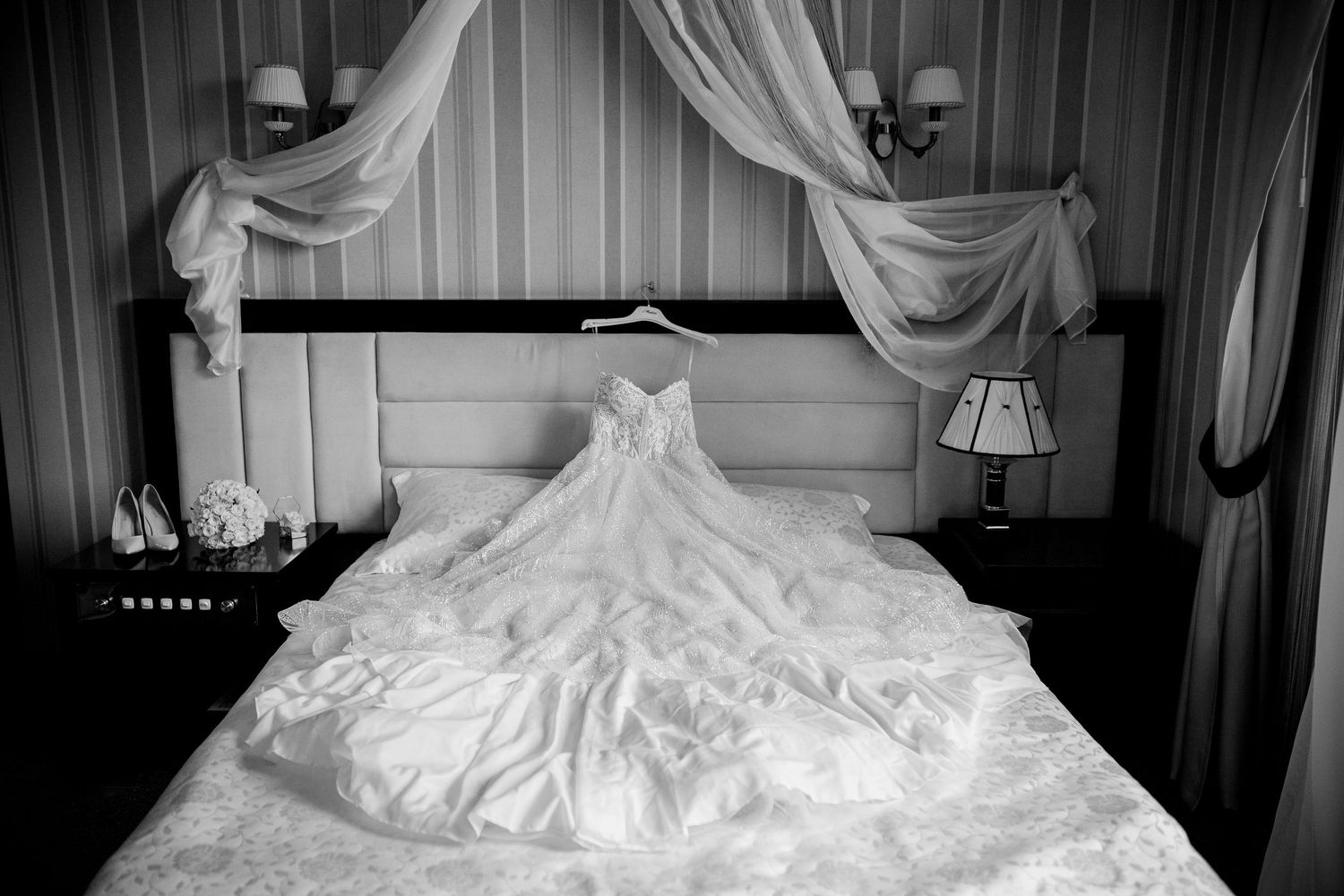 Black and white photo of an elegant wedding dress displayed on a bed in a hotel room with draped curtains.
