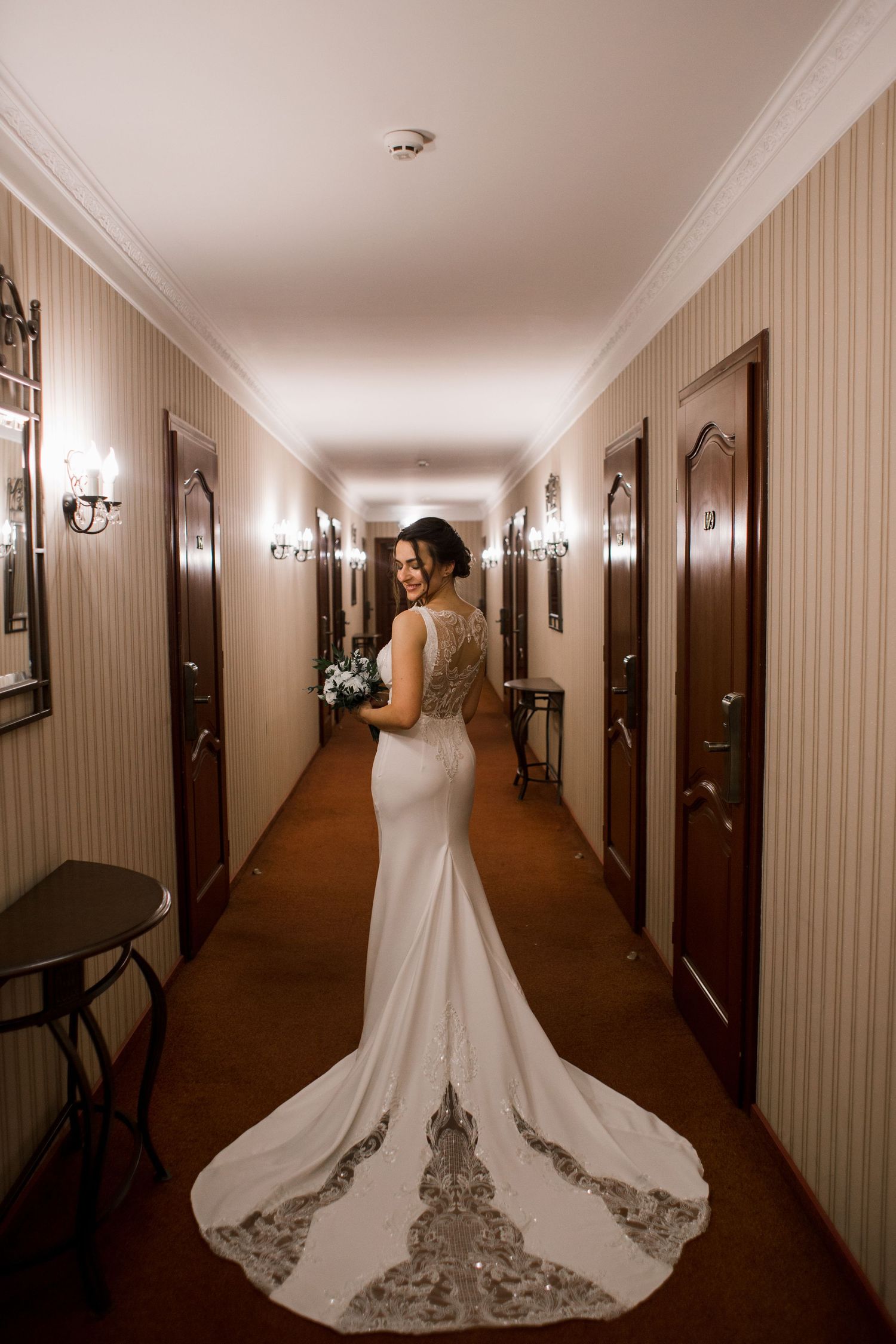Bride in elegant white mermaid gown with lace train poses in luxury hotel corridor holding bouquet.