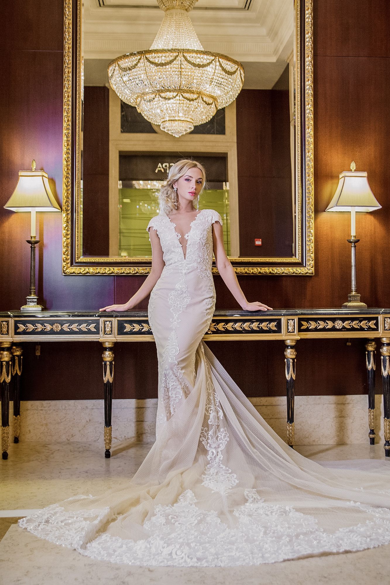 Elegant bride in lace mermaid gown with train poses in luxurious room with chandelier and ornate gold mirror.