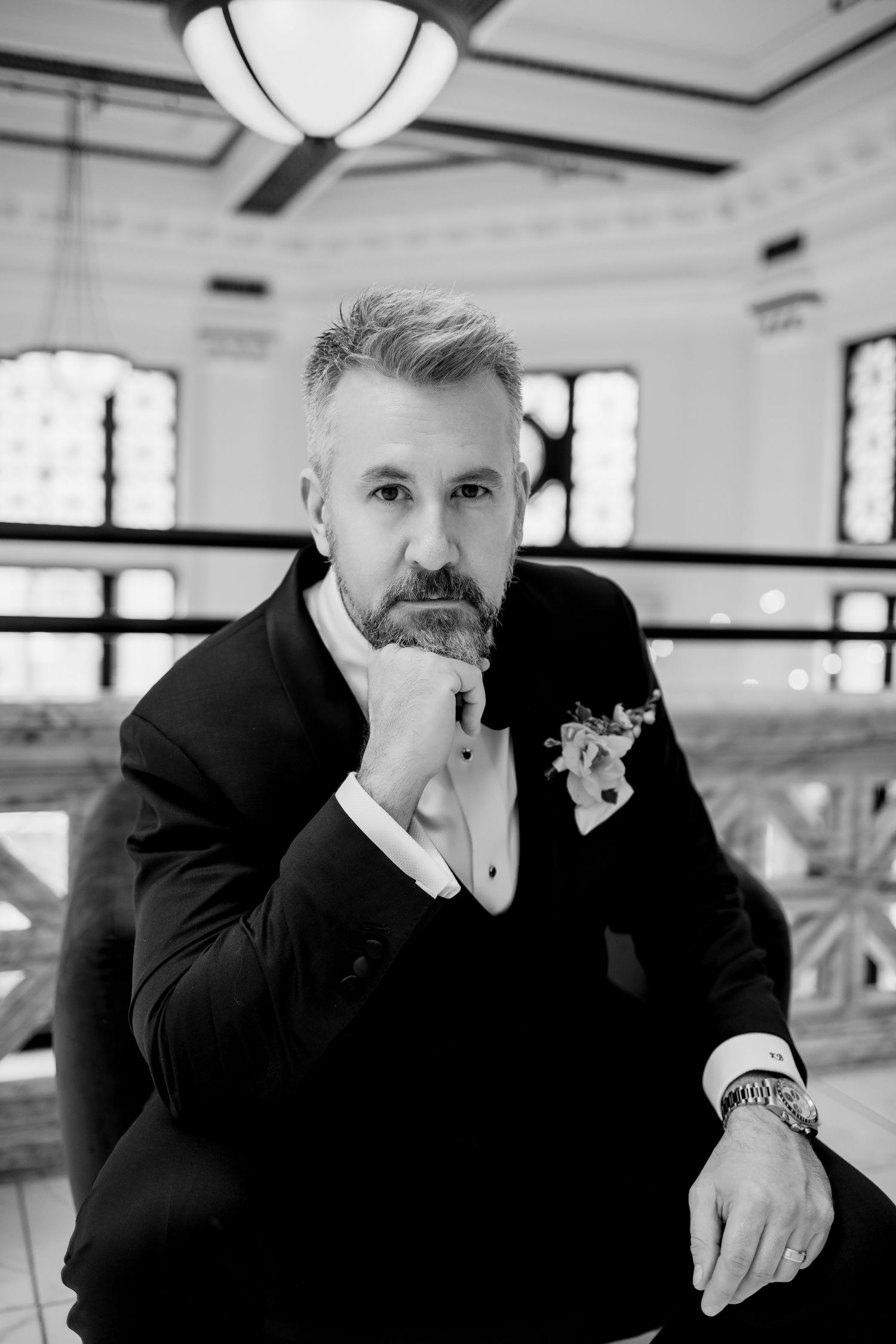 Black and white portrait of a bearded man in a suit with boutonniere, posing thoughtfully in an elegant interior.