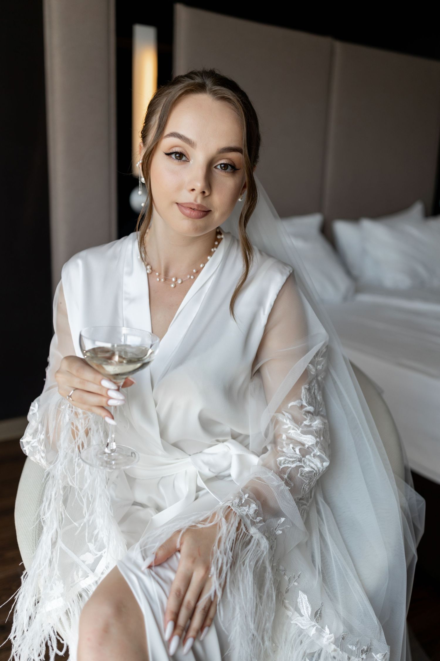 Elegant bride in white lace robe holding champagne glass, wearing pearl jewelry, sitting in luxury hotel room.