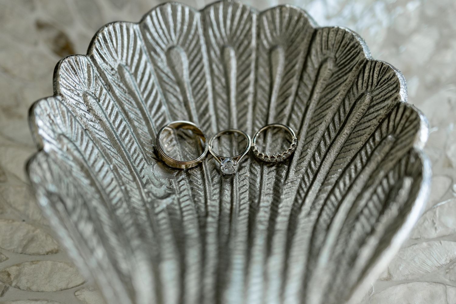 Two gold wedding rings resting in a decorative silver scallop shell dish on a marble surface.