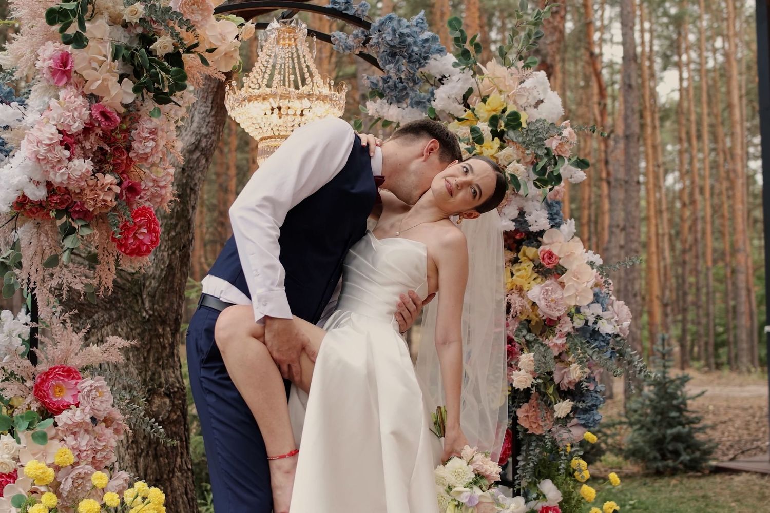Bride and groom sharing a romantic kiss under a colorful floral arch in an outdoor forest wedding ceremony.