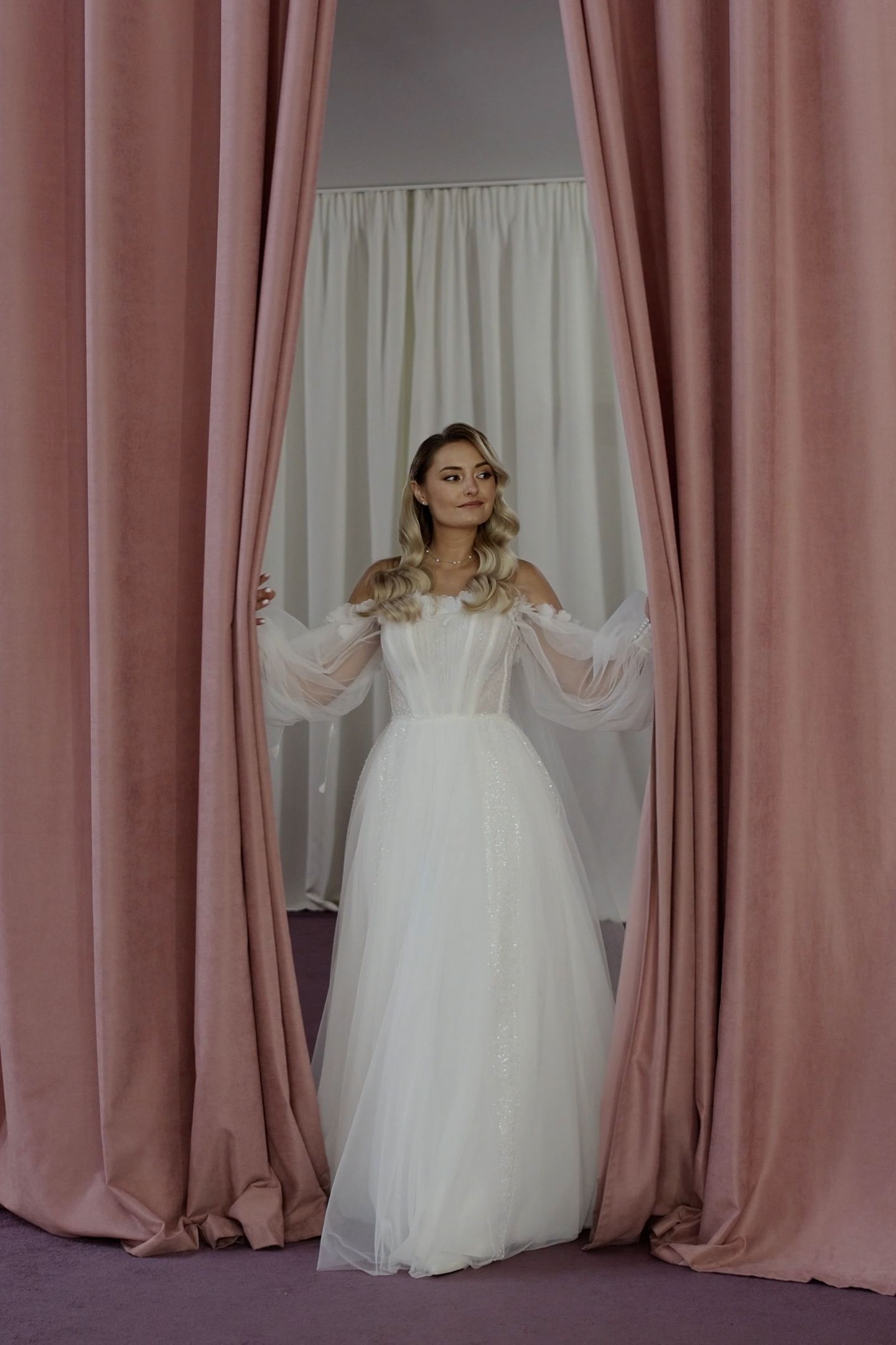 Bride in white tulle wedding gown with sheer sleeves standing between elegant dusty rose velvet curtains.