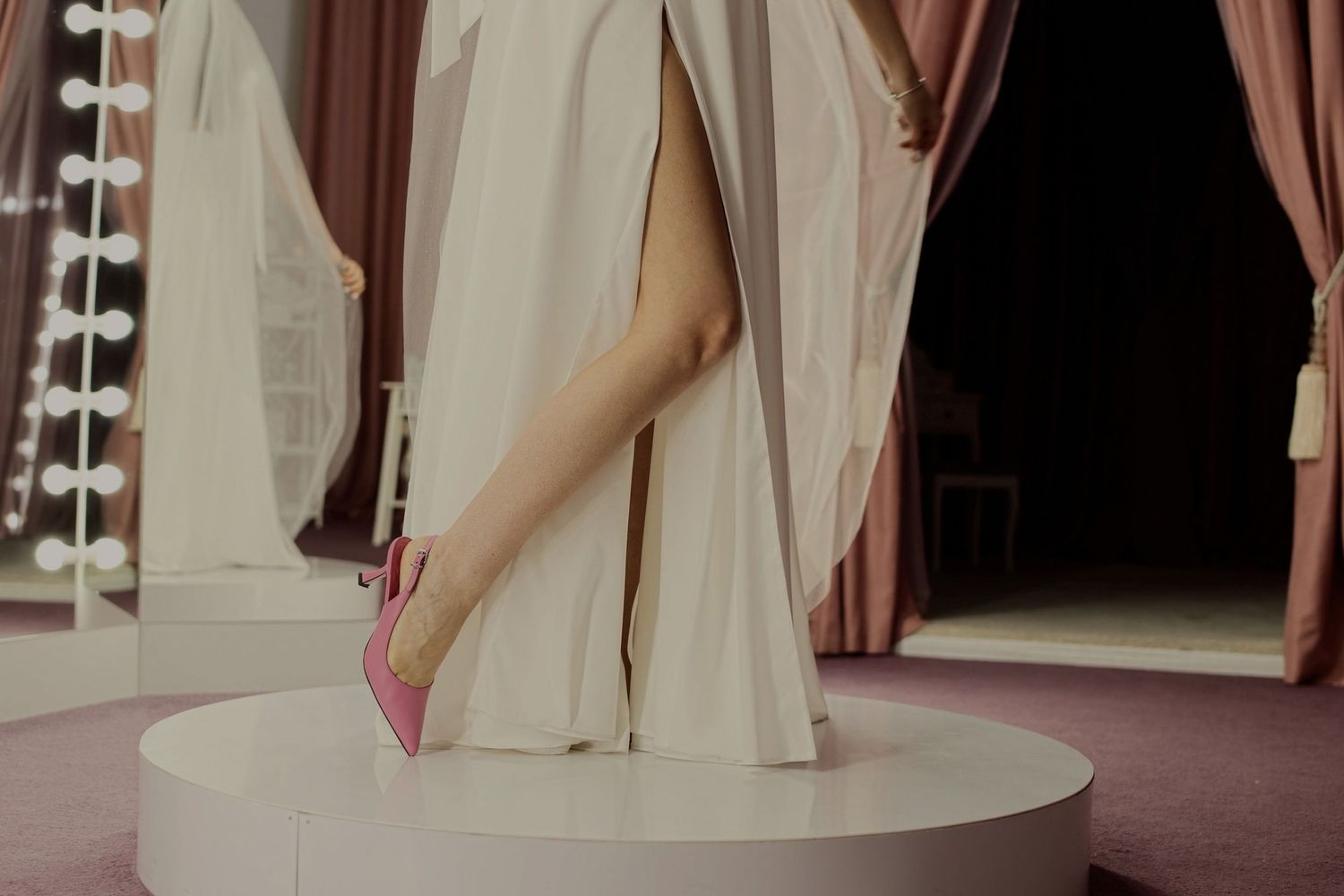Woman in white gown with thigh-high slit wearing pink heels standing on white platform near vanity mirror.