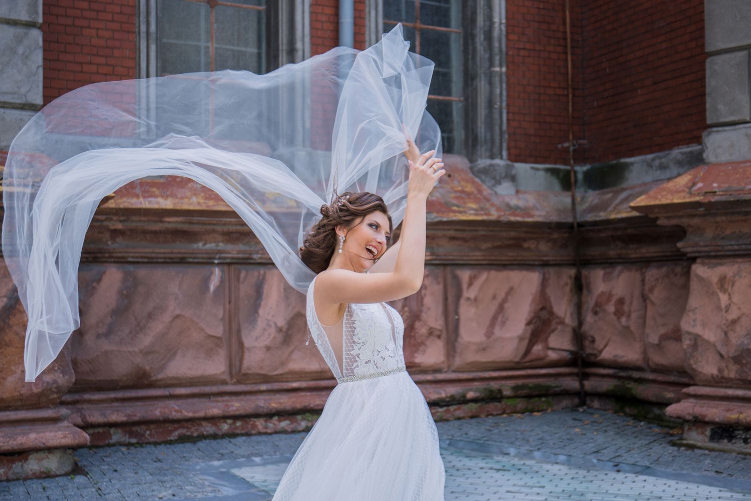 Bride in white wedding dress with flowing veil blowing in wind, posing outdoors near brick building.