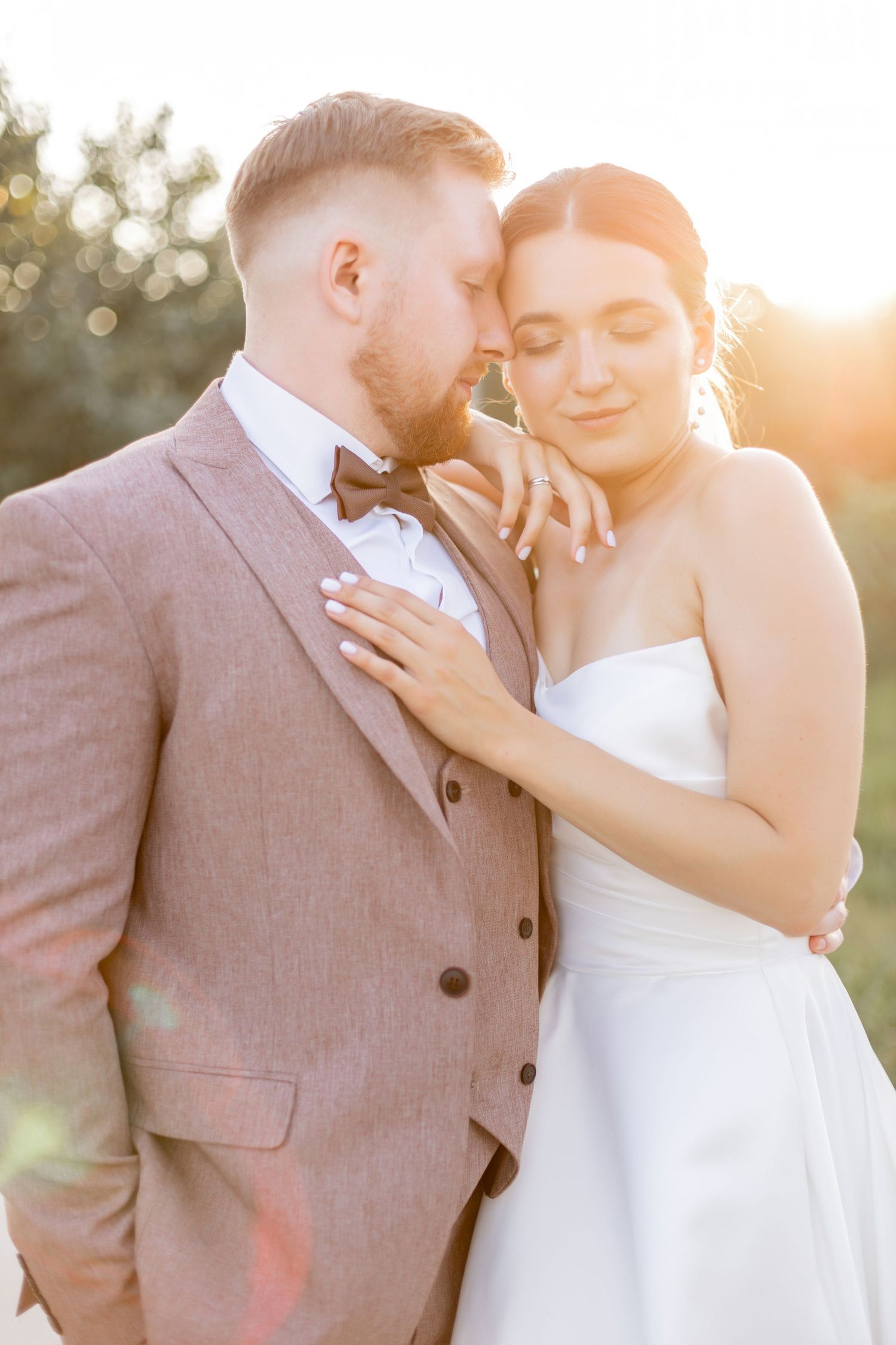 Bride and groom embrace in golden sunset light, she wears strapless white gown, he wears gray suit with bow tie.