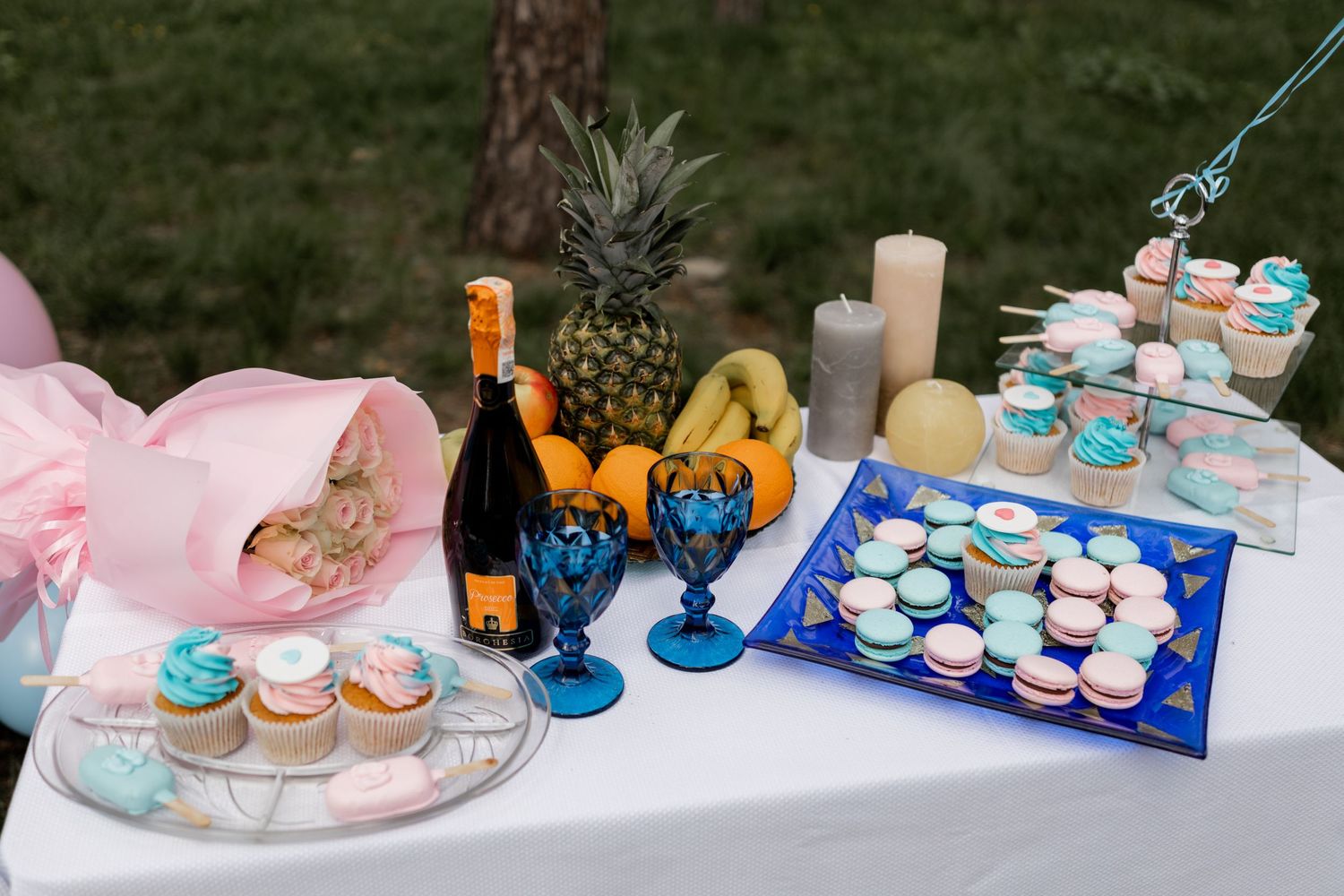 A dessert table with cupcakes, macarons, pineapple and blue goblets for a gender reveal party.