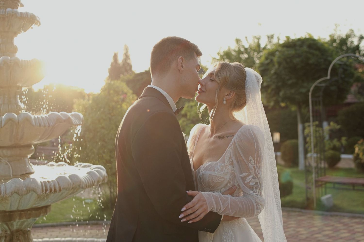 A bride and groom share a romantic moment near a fountain at golden hour, bathed in warm sunlight.