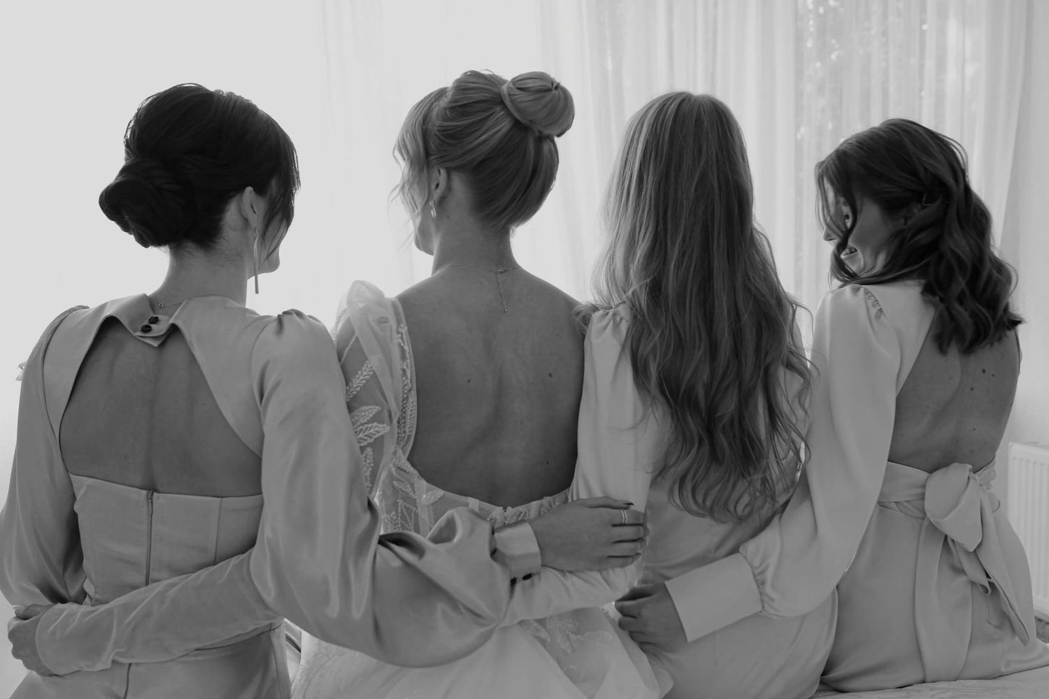Four bridesmaids in elegant dresses stand together with backs to camera in black and white wedding photo.