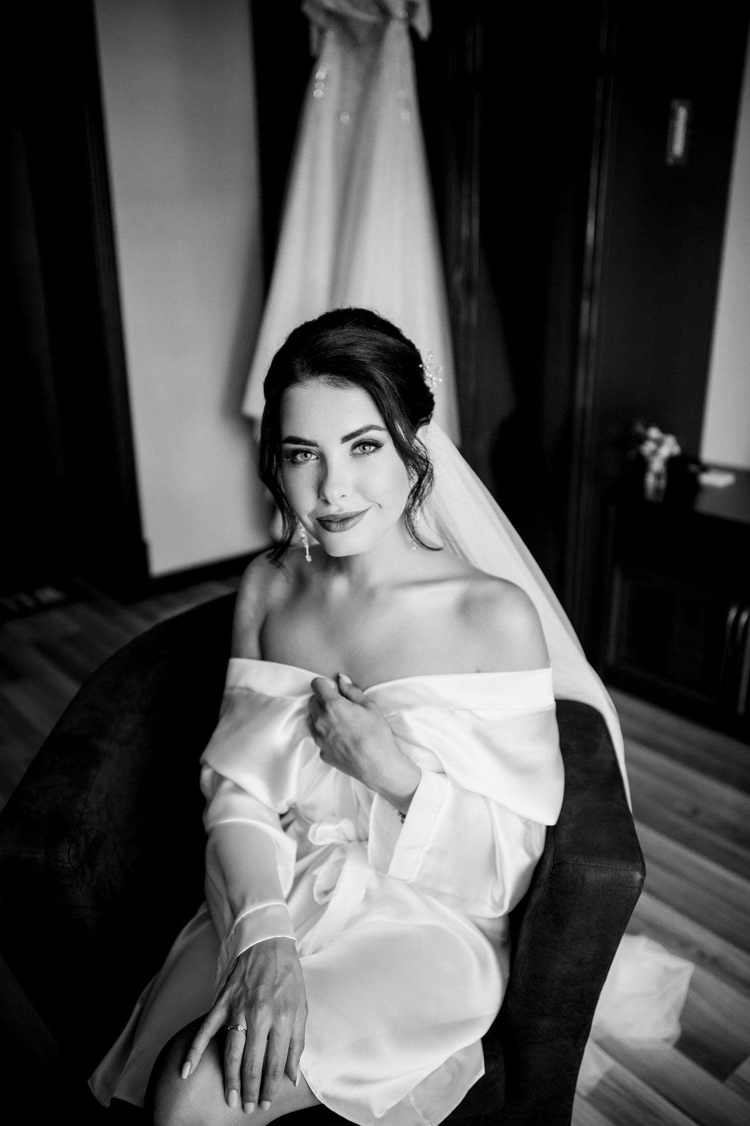Black and white portrait of a beautiful bride in a silk robe with veil, sitting elegantly before her wedding dress.
