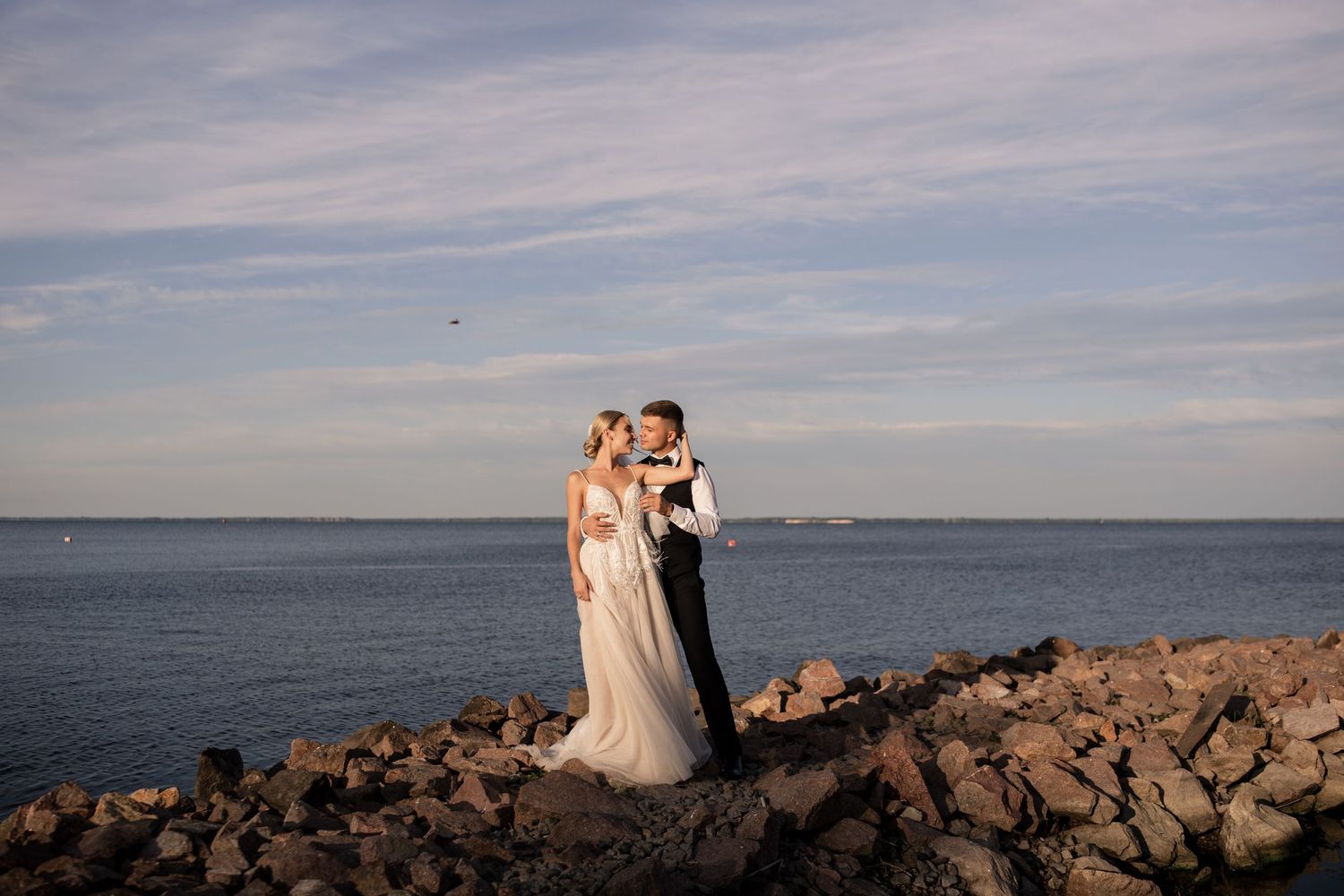 Bride and groom share a romantic kiss on rocky shoreline with ocean and blue sky backdrop at golden hour.
