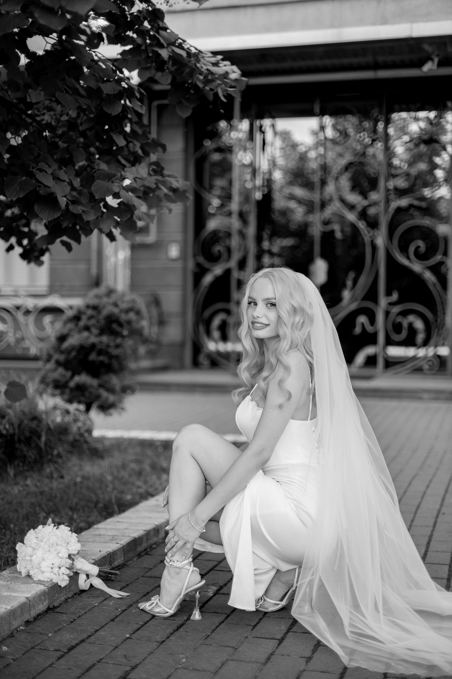 Black and white photo of a blonde bride in white gown adjusting her heels on steps, veil flowing, bouquet nearby.
