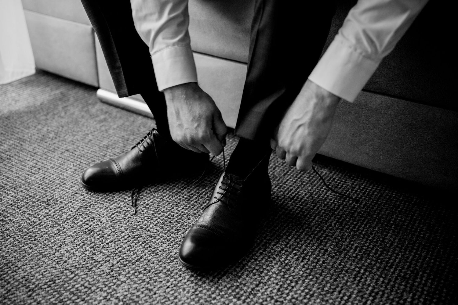 Man in formal wear tying black dress shoes on carpet floor in black and white photo.