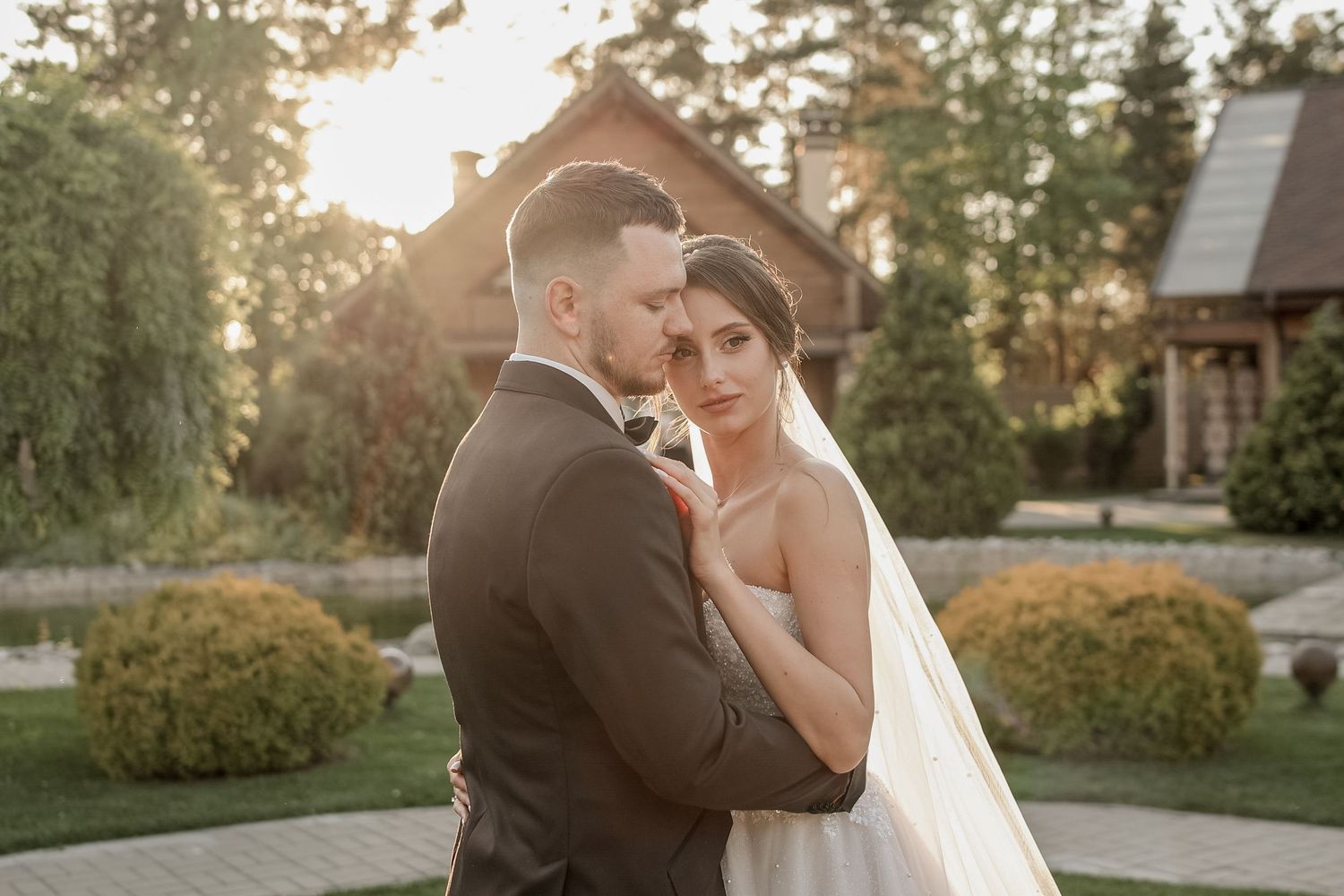 A romantic wedding couple embraces outdoors at sunset with a rustic venue and garden in the background.