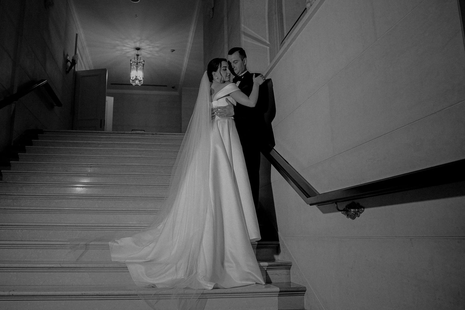 Black and white wedding photo of bride and groom embracing on grand staircase, bride in elegant gown with long veil.